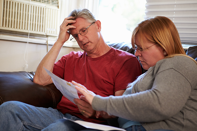 Worried Couple Sitting On Sofa Looking At Bills With Hand On Head